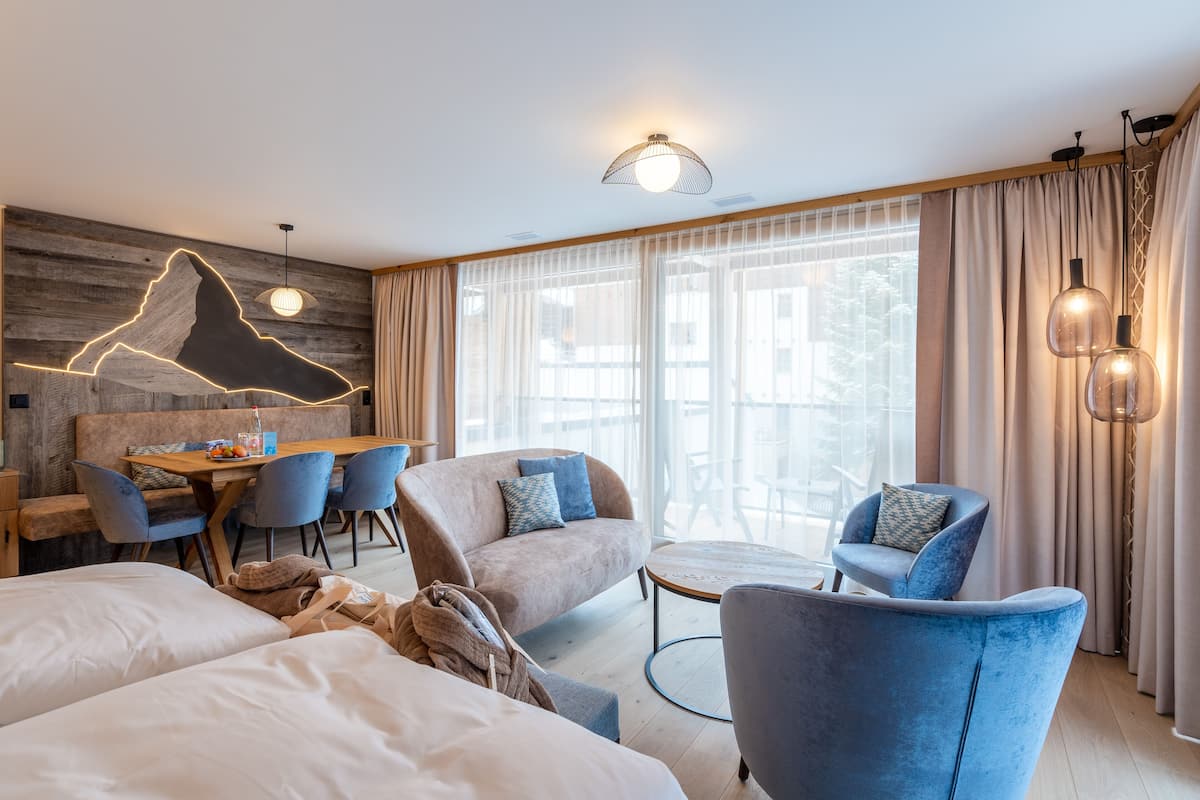Ein modernes Hotelzimmer mit einer Wand in Holzoptik, einem Essbereich, einer Couch, einem blauen Sessel und einem großen Fenster mit Vorhängen.