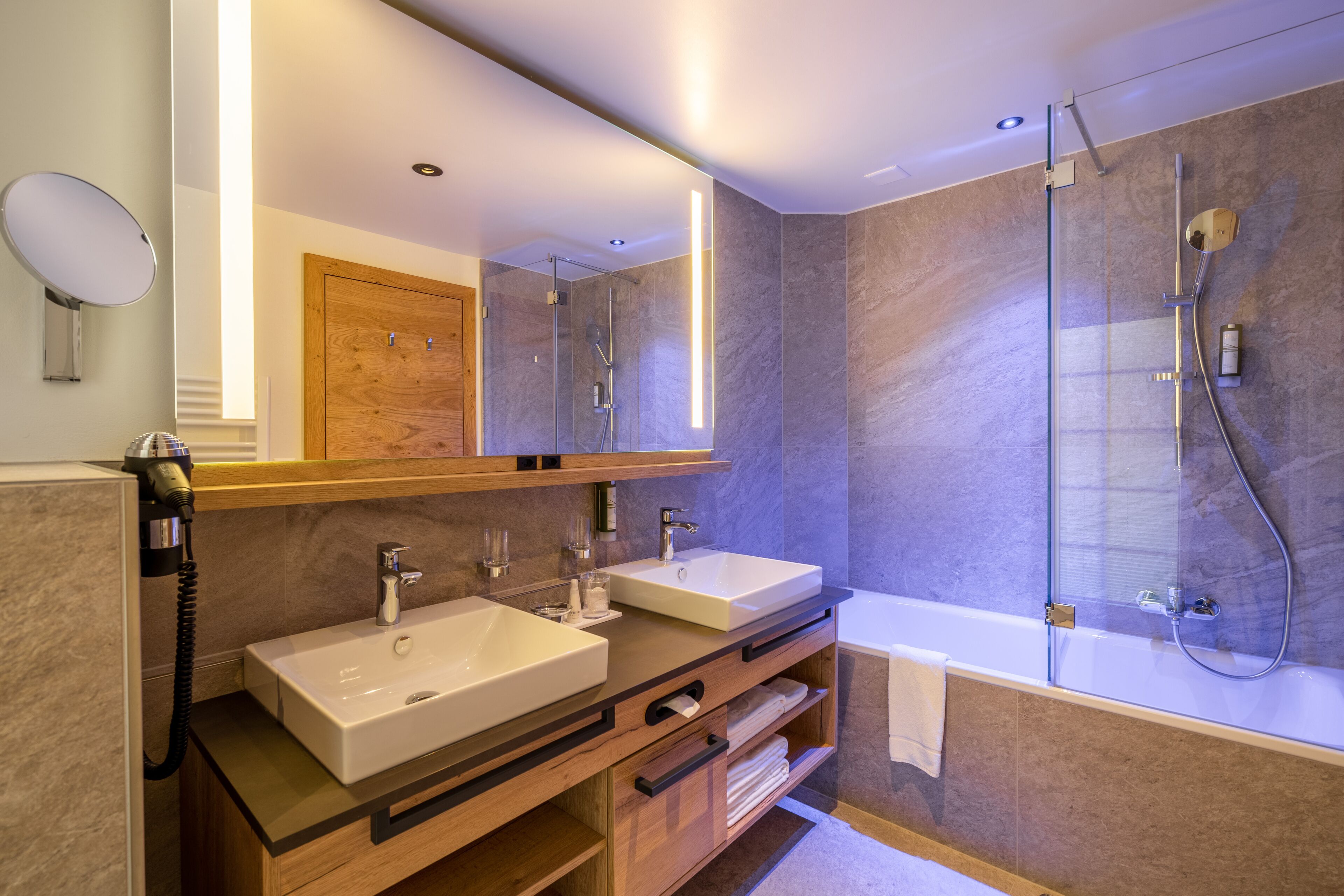 A modern bathroom with two sinks, a large mirror, a glass-enclosed shower, and a wooden vanity.