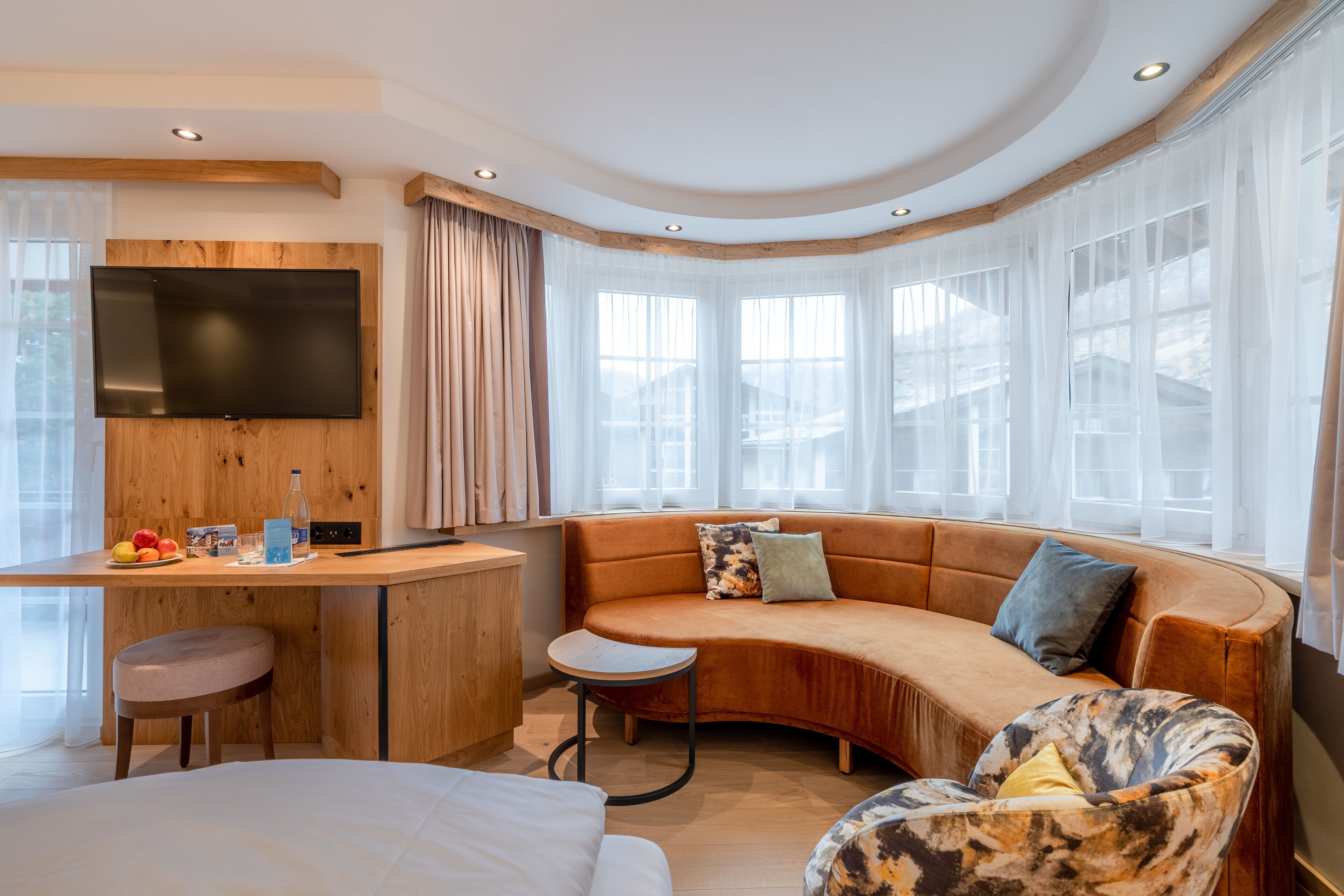 A modern hotel room with a curved sofa, a small round table, a TV mounted on a wooden panel, and large windows with sheer curtains.