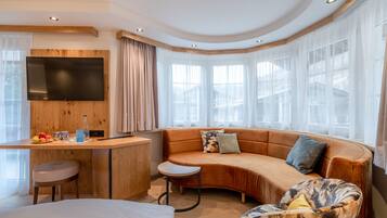 A modern hotel room with a curved sofa, a small round table, a TV mounted on a wooden panel, and large windows with sheer curtains.