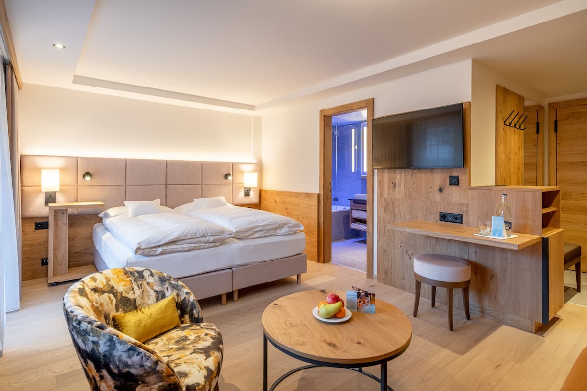 Ein modernes Hotelzimmer mit einem großen Bett, einem kleinen Schreibtisch, einem Stuhl und einem runden Tisch mit Obst.