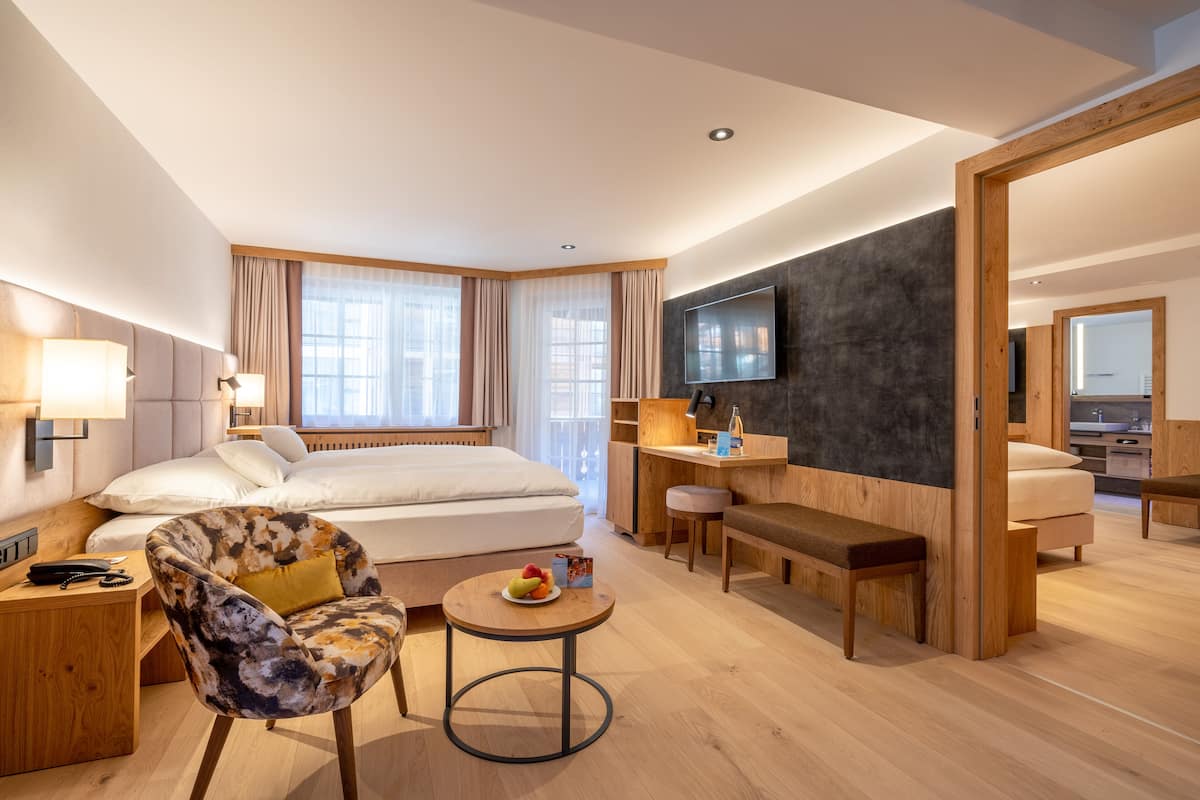 Ein modernes Hotelzimmer mit einem großen Bett, einem Schreibtisch, einem Sessel, einem runden Tisch mit Früchten und einem an der Wand befestigten Fernseher.