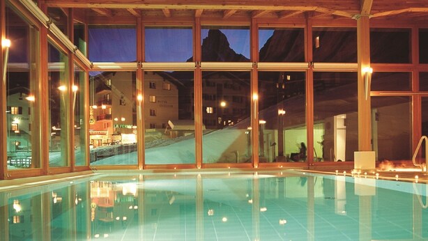 Una piscina interior con amplios ventanales, vigas de madera y vistas a una montaña y edificios.