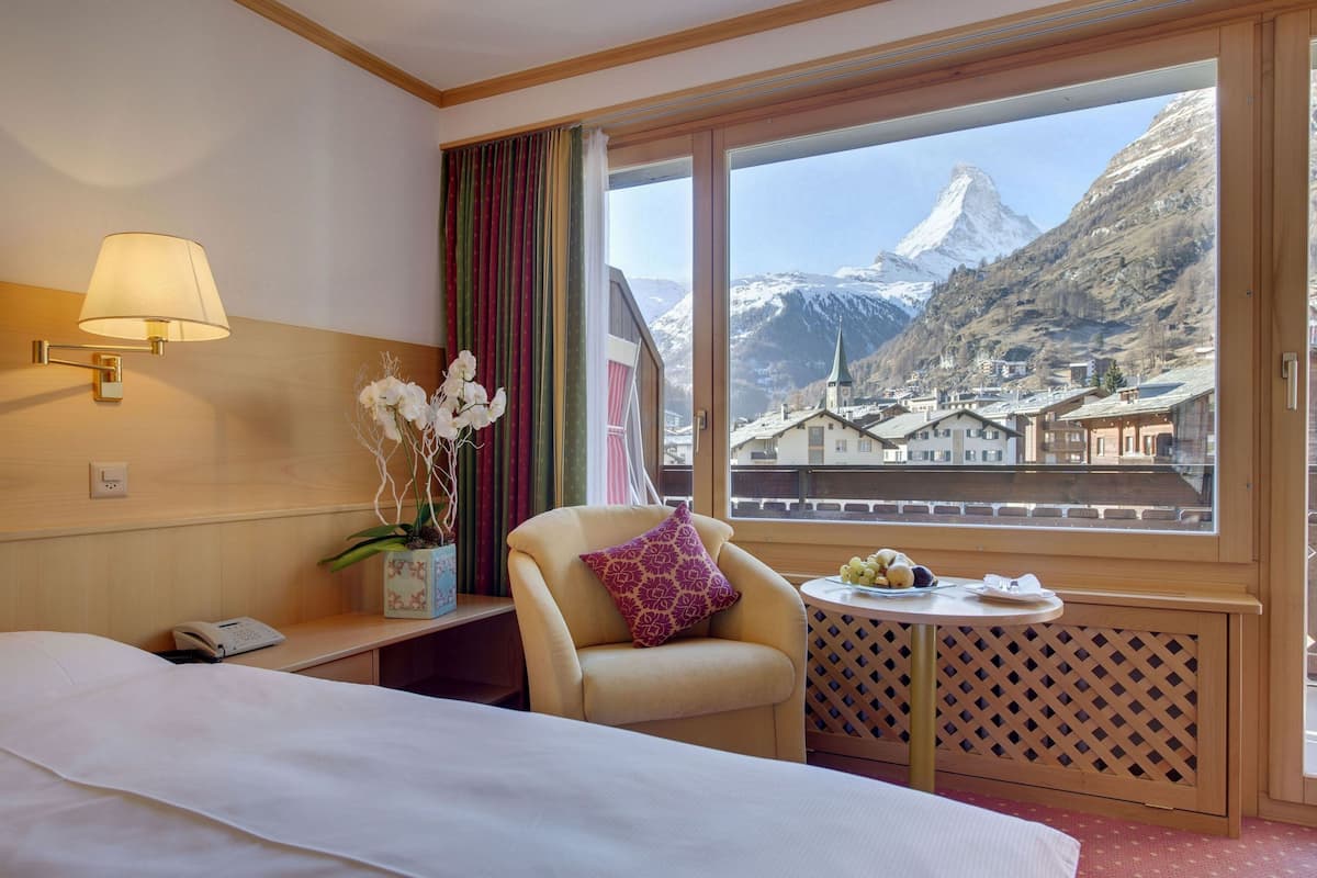 A hotel room with a bed, a chair, a small table with fruit, a lamp, and a view of a mountain.