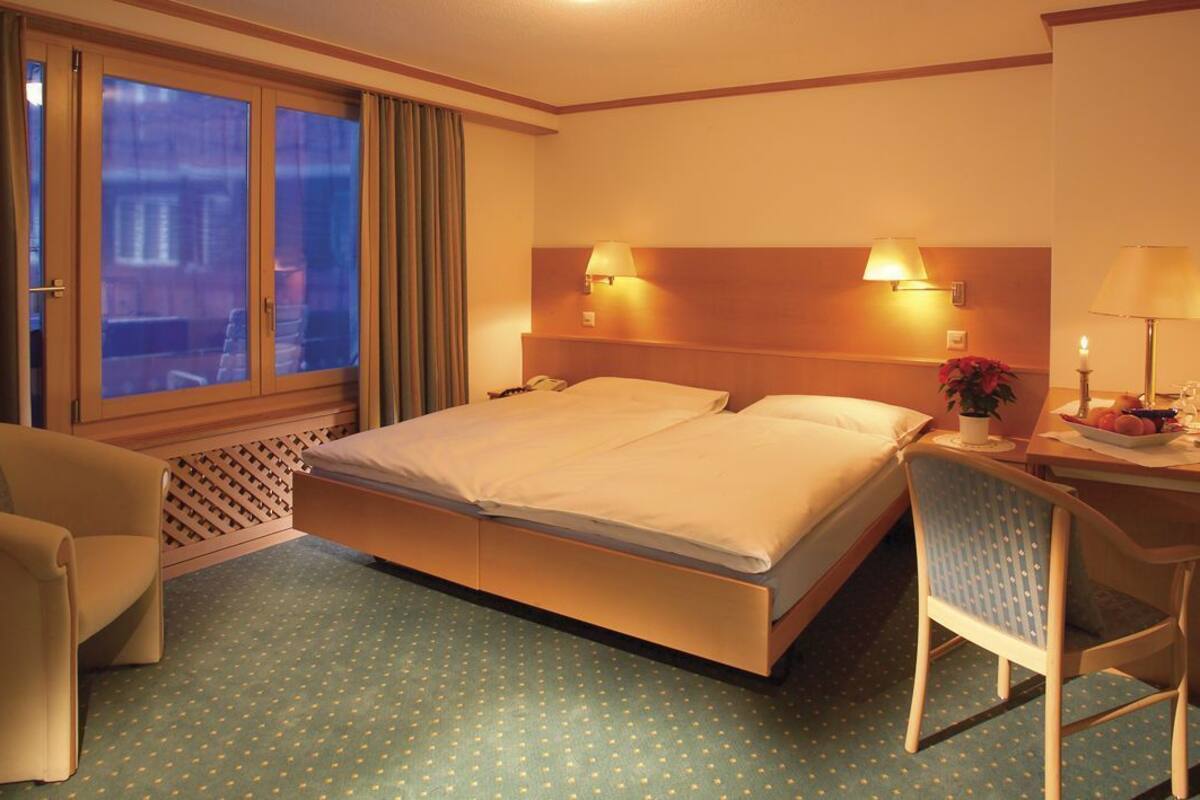 A hotel room with a bed, a desk with a chair, a lamp, and a window with curtains.