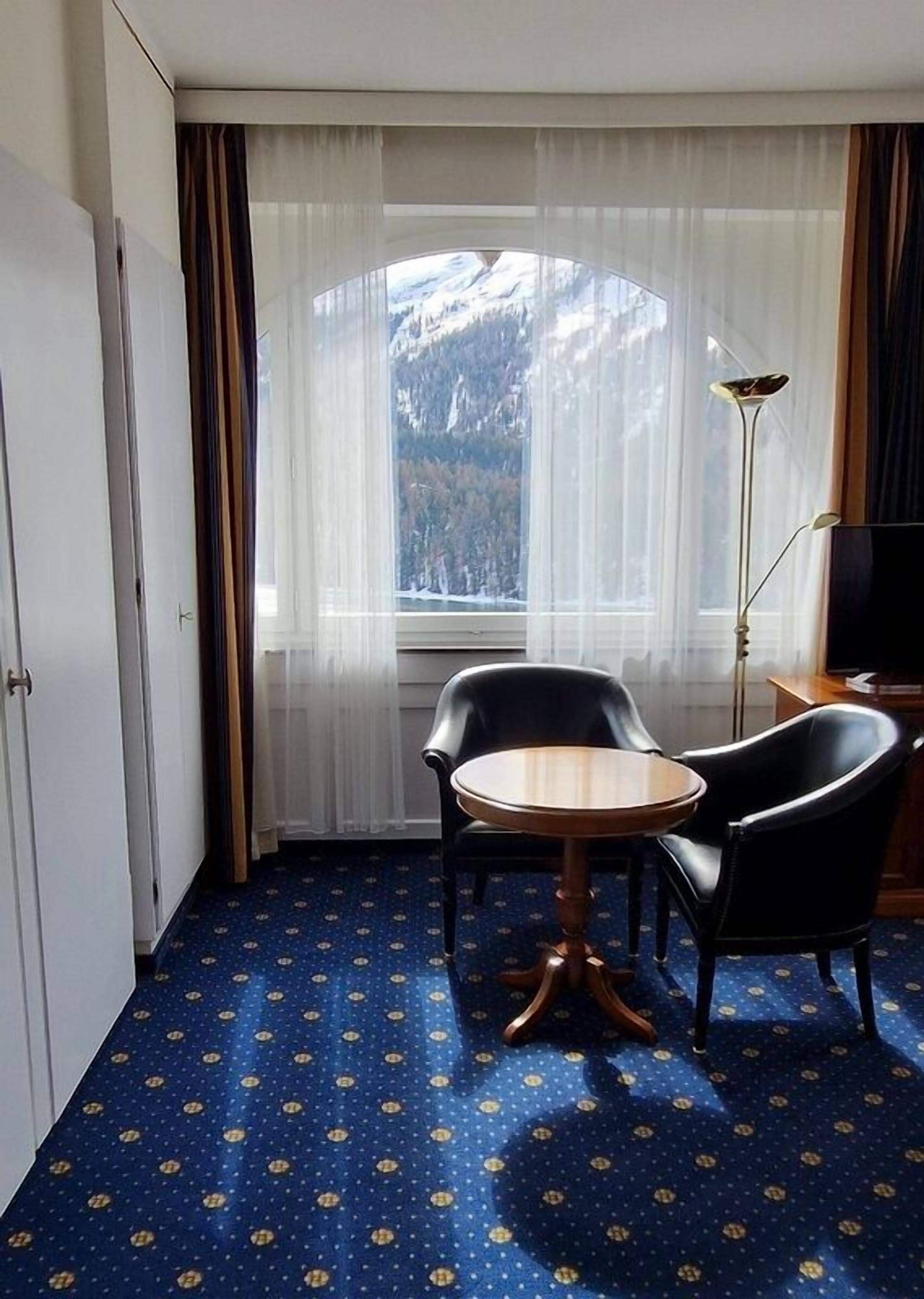 A hotel room with a round wooden table, two black chairs, a floor lamp, and a window with a mountain view.