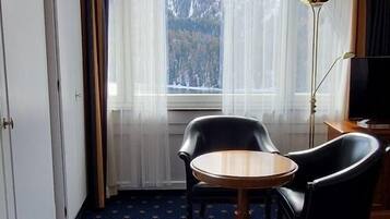 A hotel room with a round wooden table, two black chairs, a floor lamp, and a window with a mountain view.
