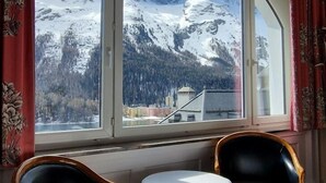 Ein Zimmer mit einem großen Fenster, das einen Blick auf schneebedeckte Berge und eine darunterliegende Stadt bietet. Im Raum befinden sich zwei Stühle und ein kleiner Tisch.