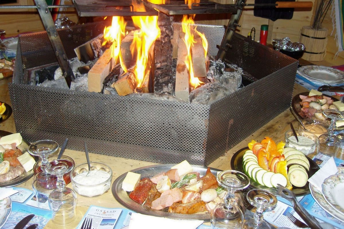 A metal grill with a fire burning inside, surrounded by various food items and utensils on a table.