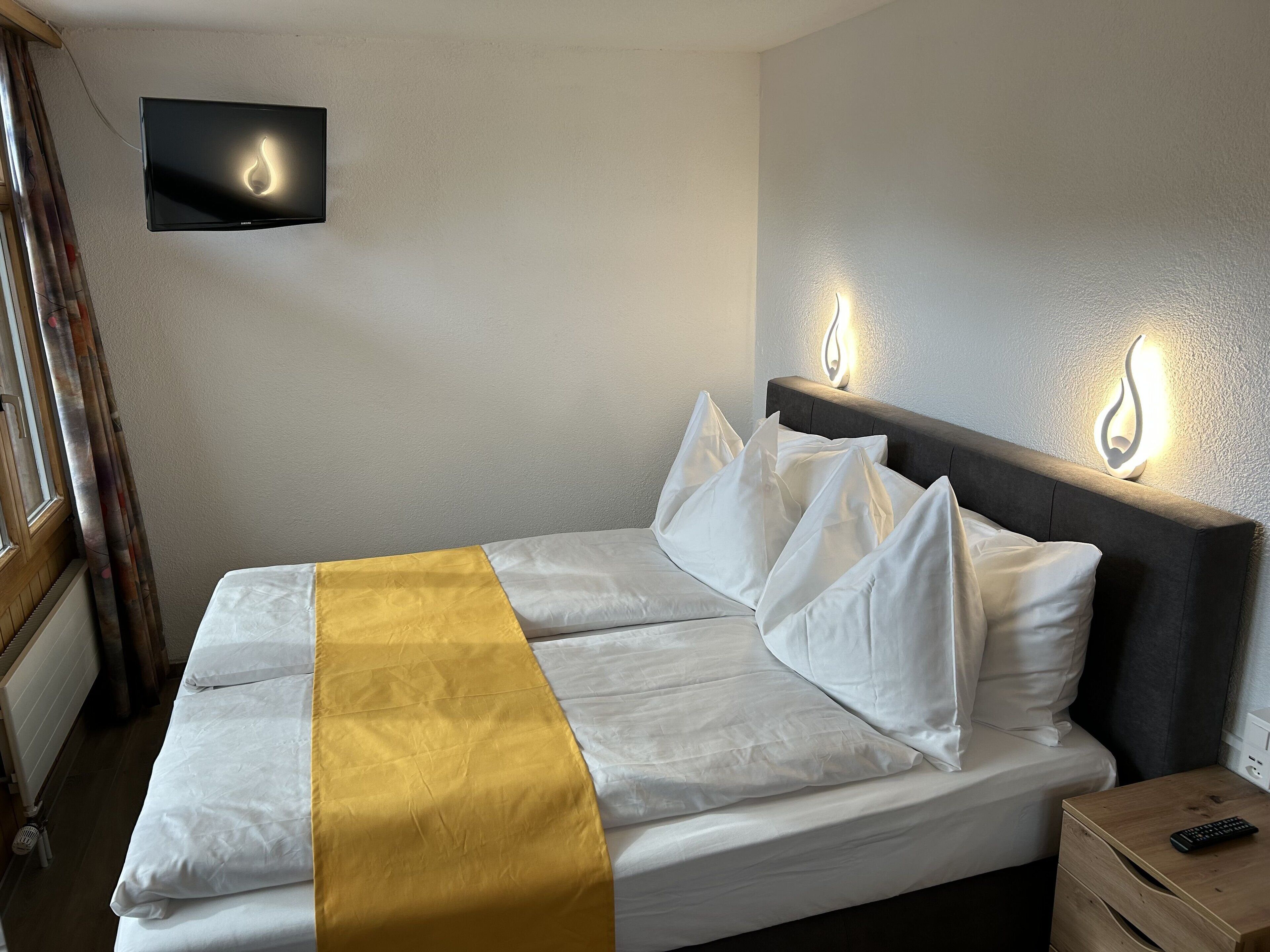 A neatly made bed with white and yellow bedding, a flat-screen TV mounted on the wall, and bedside lamps.