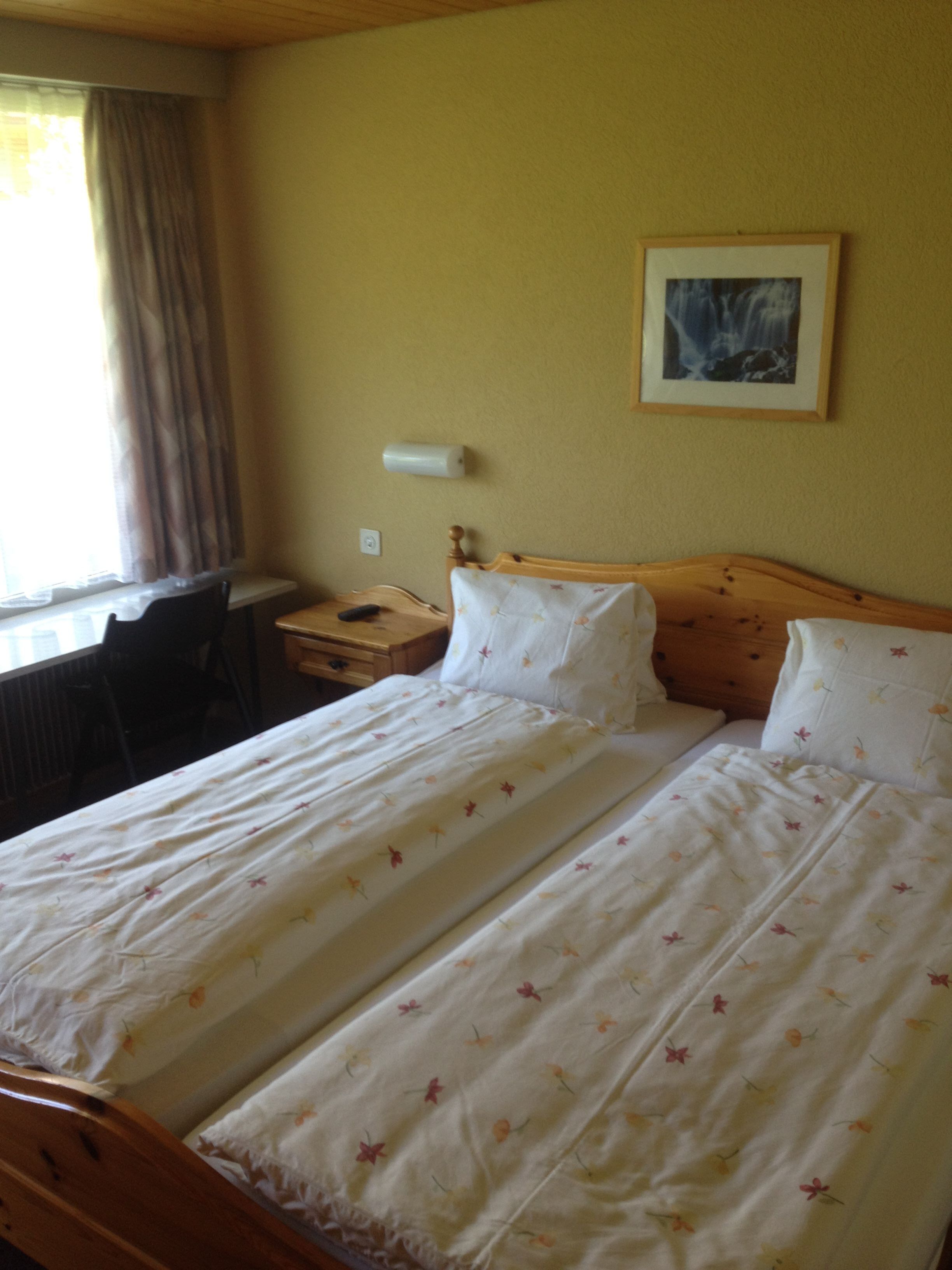 A double bed with floral bedding, a wooden headboard, a bedside table with a lamp, and a window with curtains.