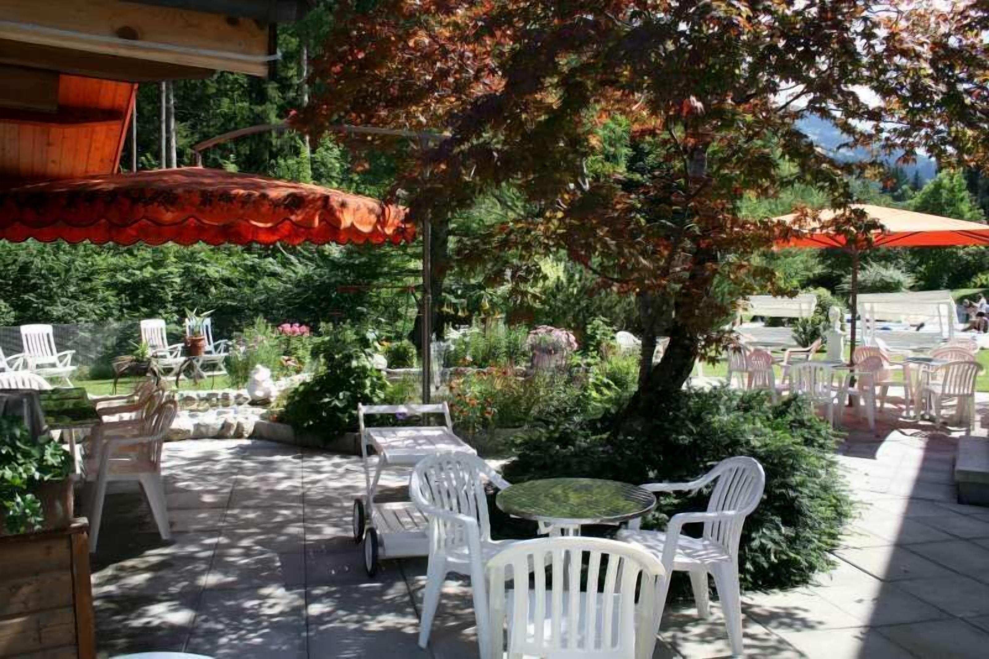 An outdoor patio area with white plastic chairs and tables, surrounded by greenery and trees.