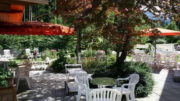 An outdoor patio area with white plastic chairs and tables, surrounded by greenery and trees.