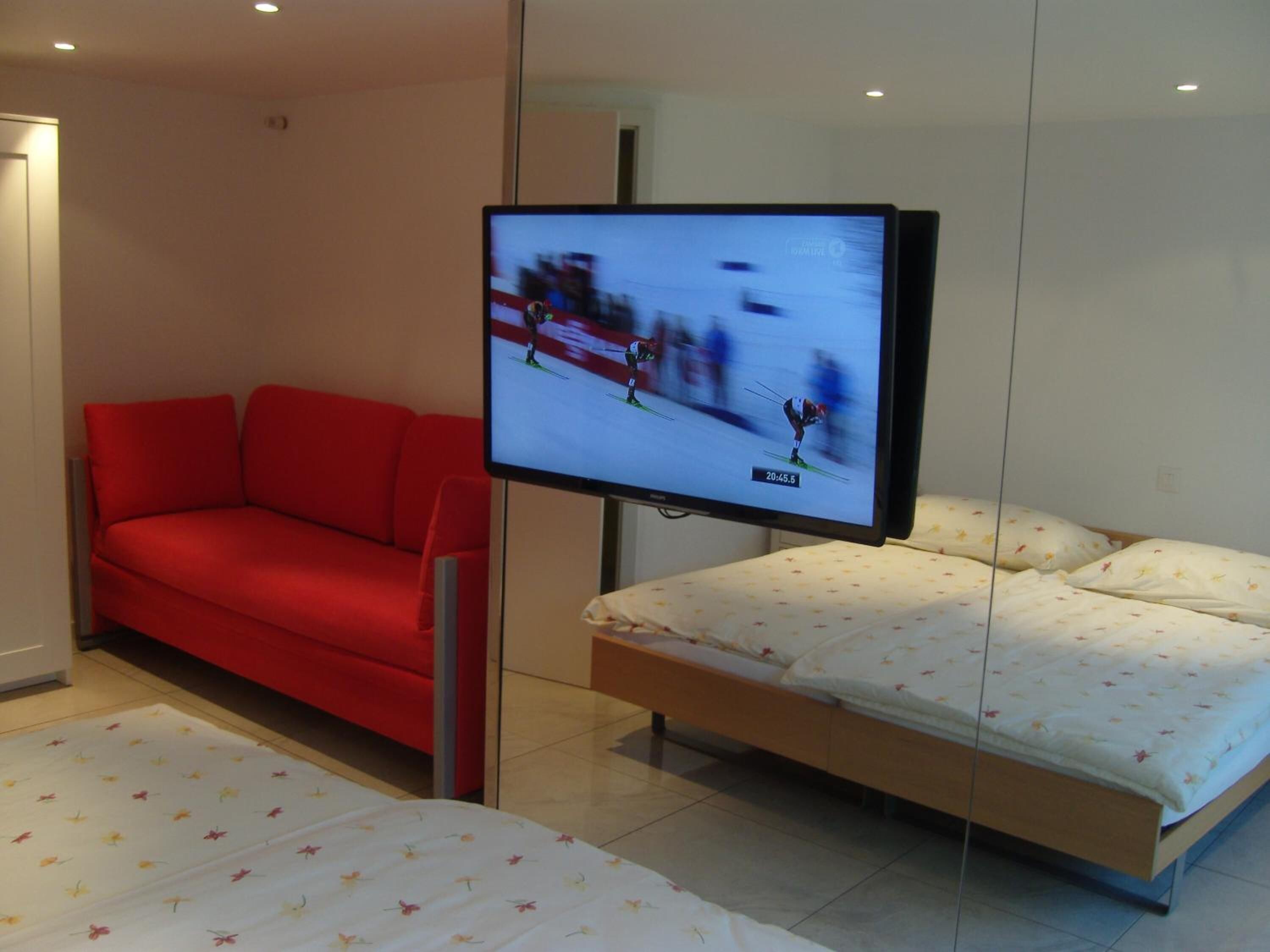 Una habitación de hotel moderna con un sofá rojo, una cama con ropa de cama floral y un televisor mostrando un evento deportivo borroso.