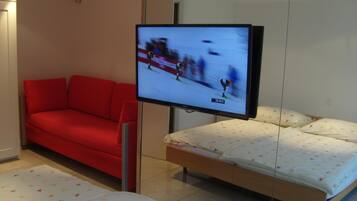 Una habitación de hotel moderna con un sofá rojo, una cama con ropa de cama floral y un televisor mostrando un evento deportivo borroso.