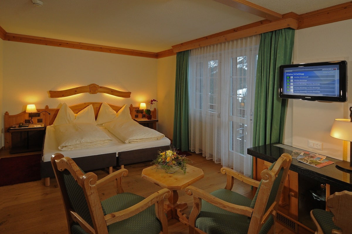 A hotel room with a bed, two chairs, a small table with a flower vase, a TV on the wall, and a window with curtains.