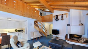 A spacious living area with a wooden staircase, a fireplace, a television, and a kitchen in the background. - Hotel Kirchbuehl (Grindelwald)