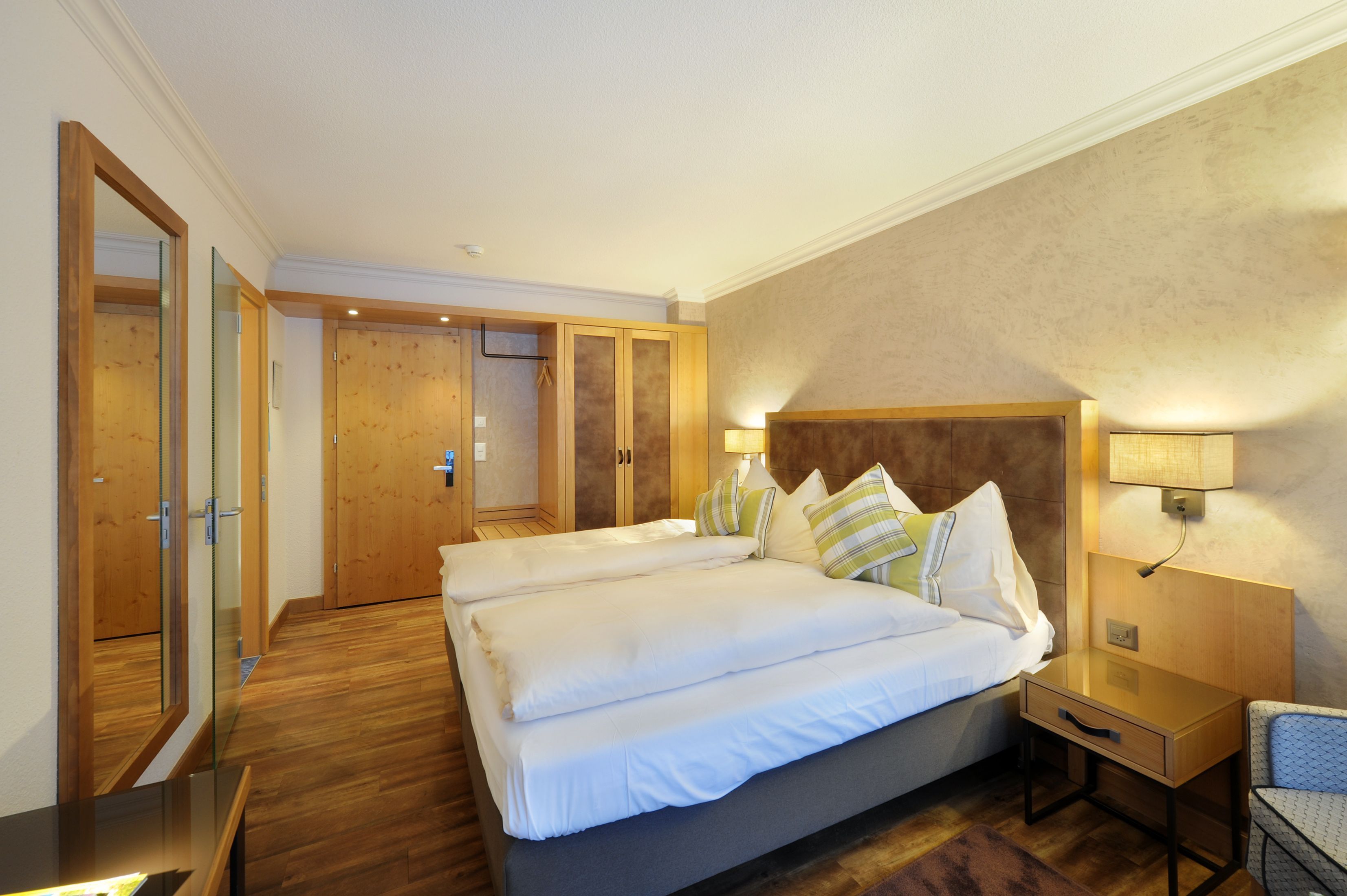 A hotel room with a large bed, wooden flooring, a nightstand, a lamp, and a chair.
