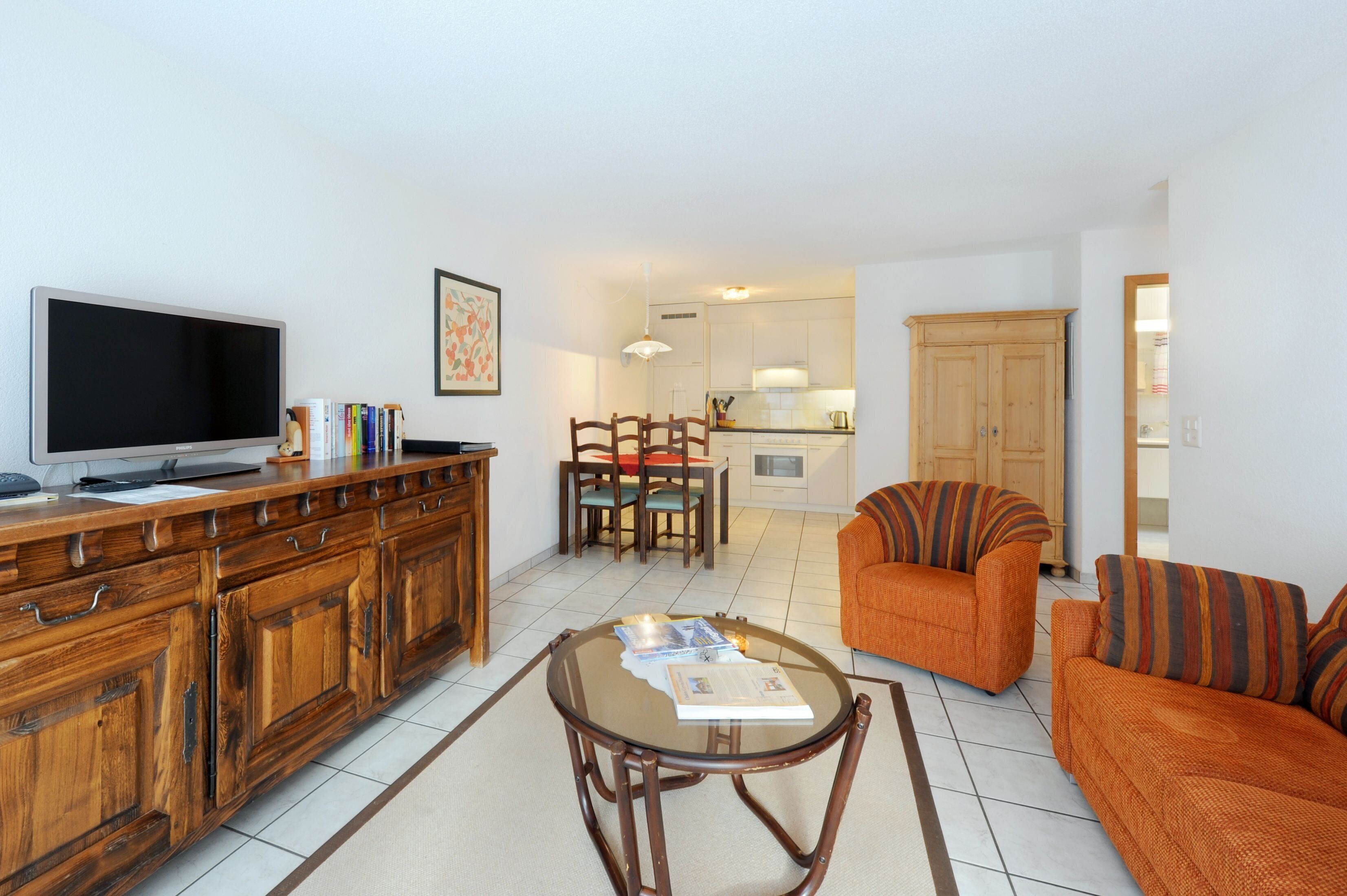A living room with a TV, wooden cabinet, orange sofa, and a dining area with chairs and a table.