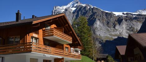 A wooden chalet with a balcony, a mountain backdrop, and a clear sky.