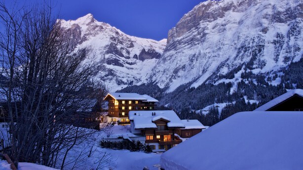 A snow-covered mountain lodge with multiple windows and a well-lit exterior, surrounded by snow-covered trees and mountains. - Hotel Kirchbuehl (Grindelwald)