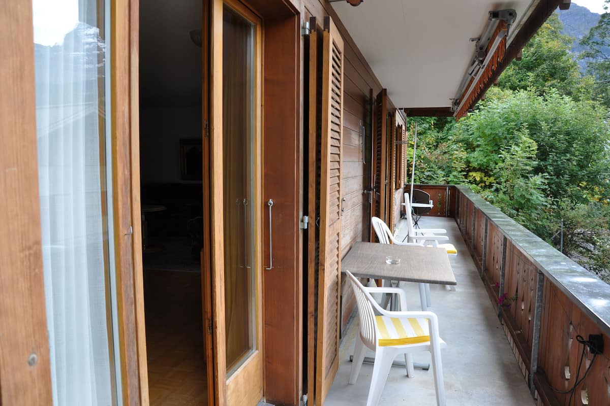 Une porte en bois ouverte donne sur un balcon où se trouvent une table et des chaises, et d’où l’on peut admirer un paysage verdoyant et luxuriant.