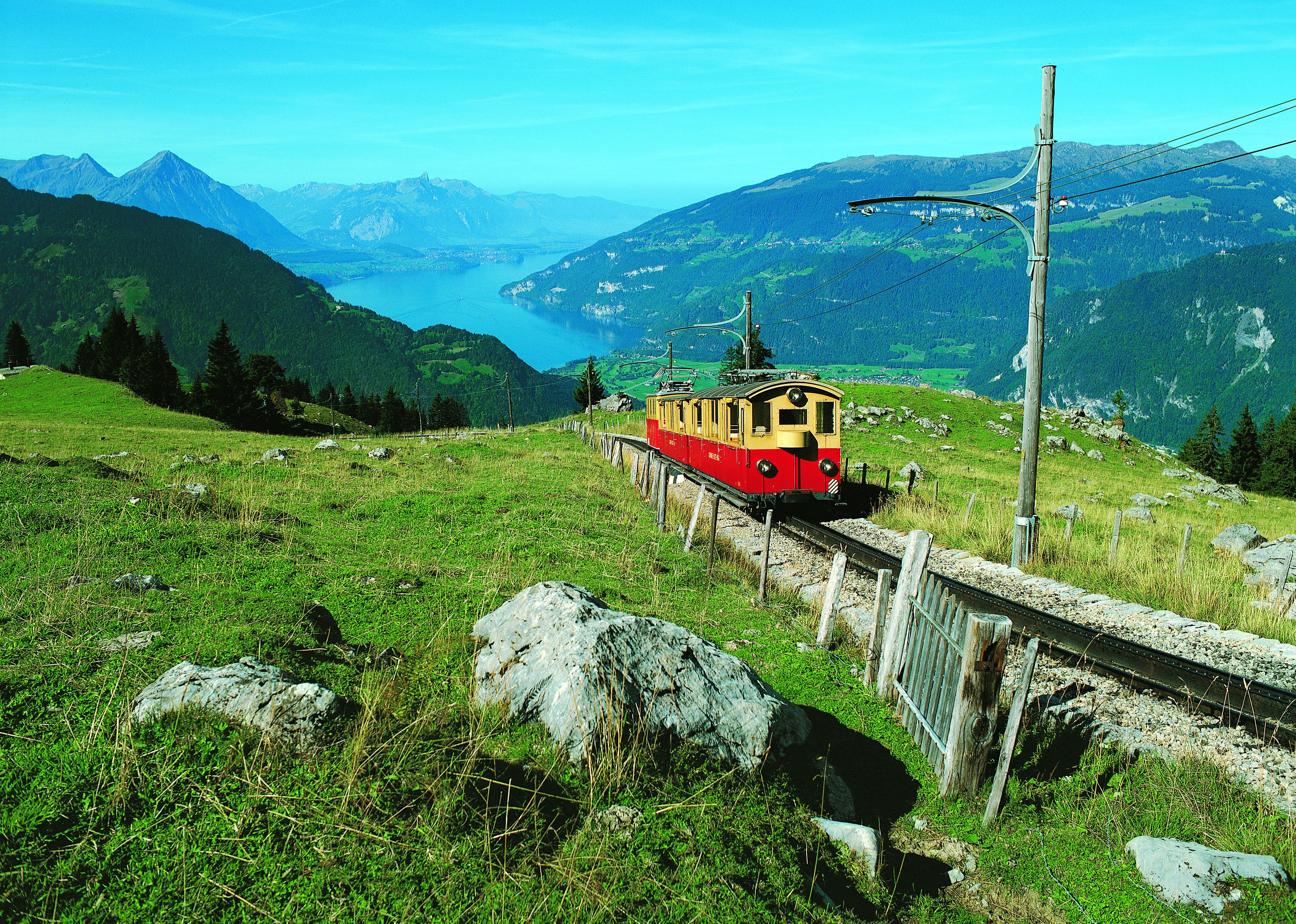 A red and yellow train on a mountain track with a scenic view of mountains and a lake.