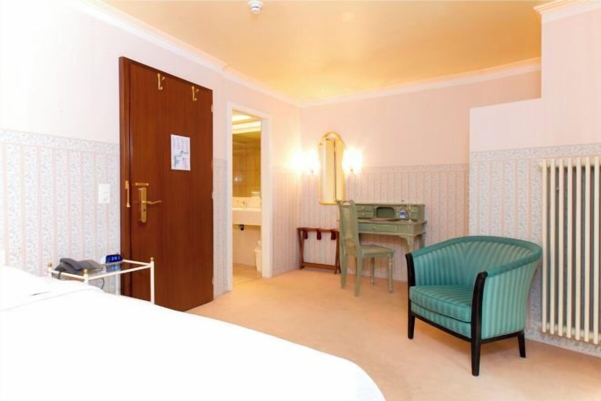 Habitación de hotel con una puerta de madera, un sillón verde, un escritorio pequeño con espejo y un radiador.
