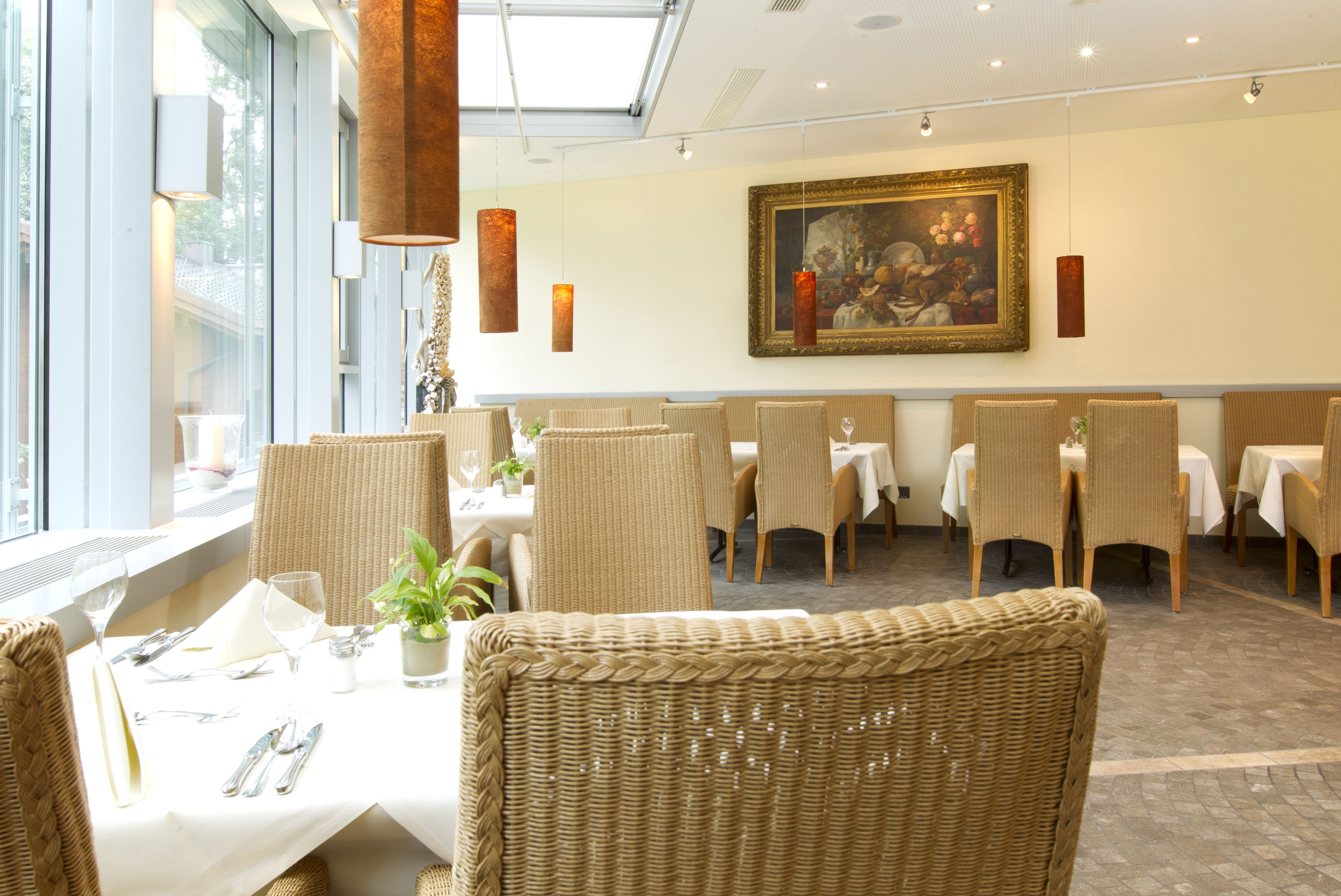 A restaurant with wicker chairs, white tablecloths, and a large painting on the wall.