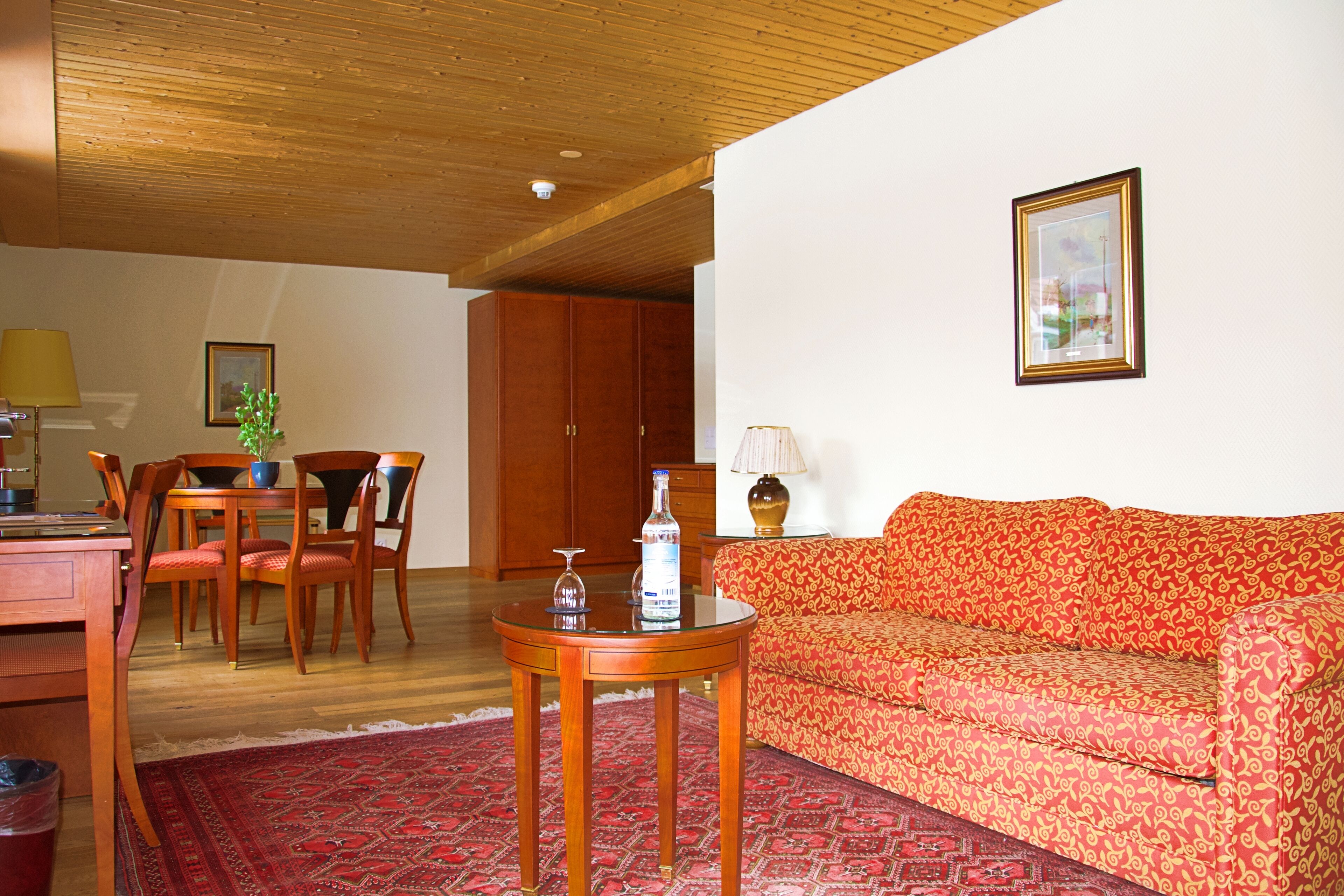 A living room with a wooden ceiling, a red patterned sofa, a wooden dining table with chairs, and a red patterned rug.