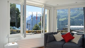 A room with a grey sofa, a small white table, and large windows offering a view of a mountain landscape and a train station. - Hotel Silberhorn - Residences & Spa Wengen (Lauterbrunnen)