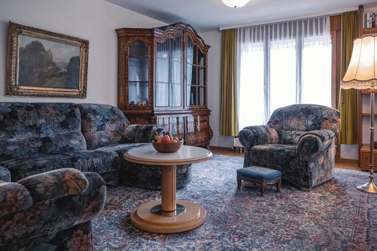 Un salon avec un canapé à motifs, une table basse ronde, une table d’appoint sur laquelle se trouve un saladier rempli de fruits, un meuble en bois et une lampe.