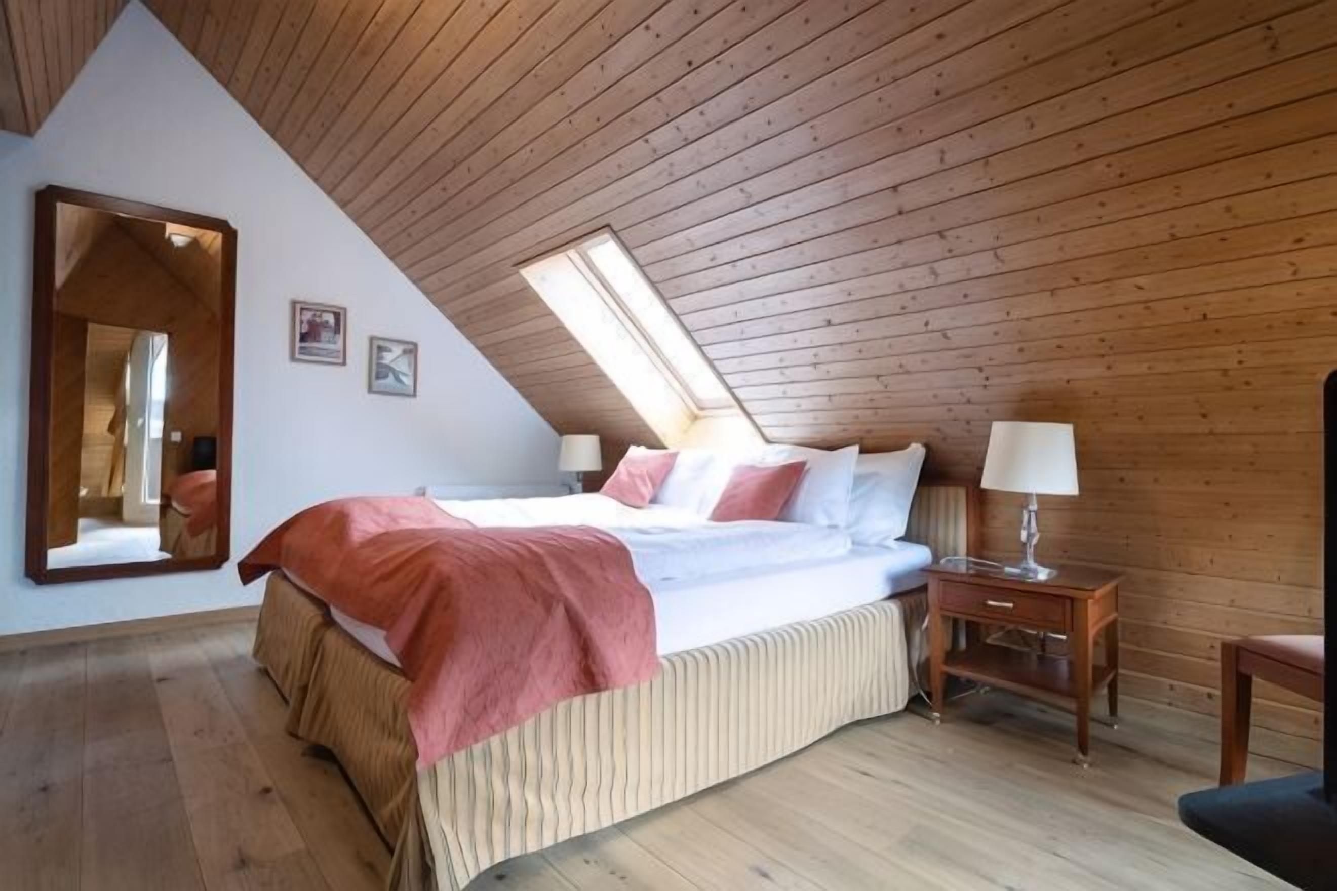 A bedroom with a sloped ceiling, a bed with a red blanket, two bedside tables with lamps, and a large mirror on the wall.