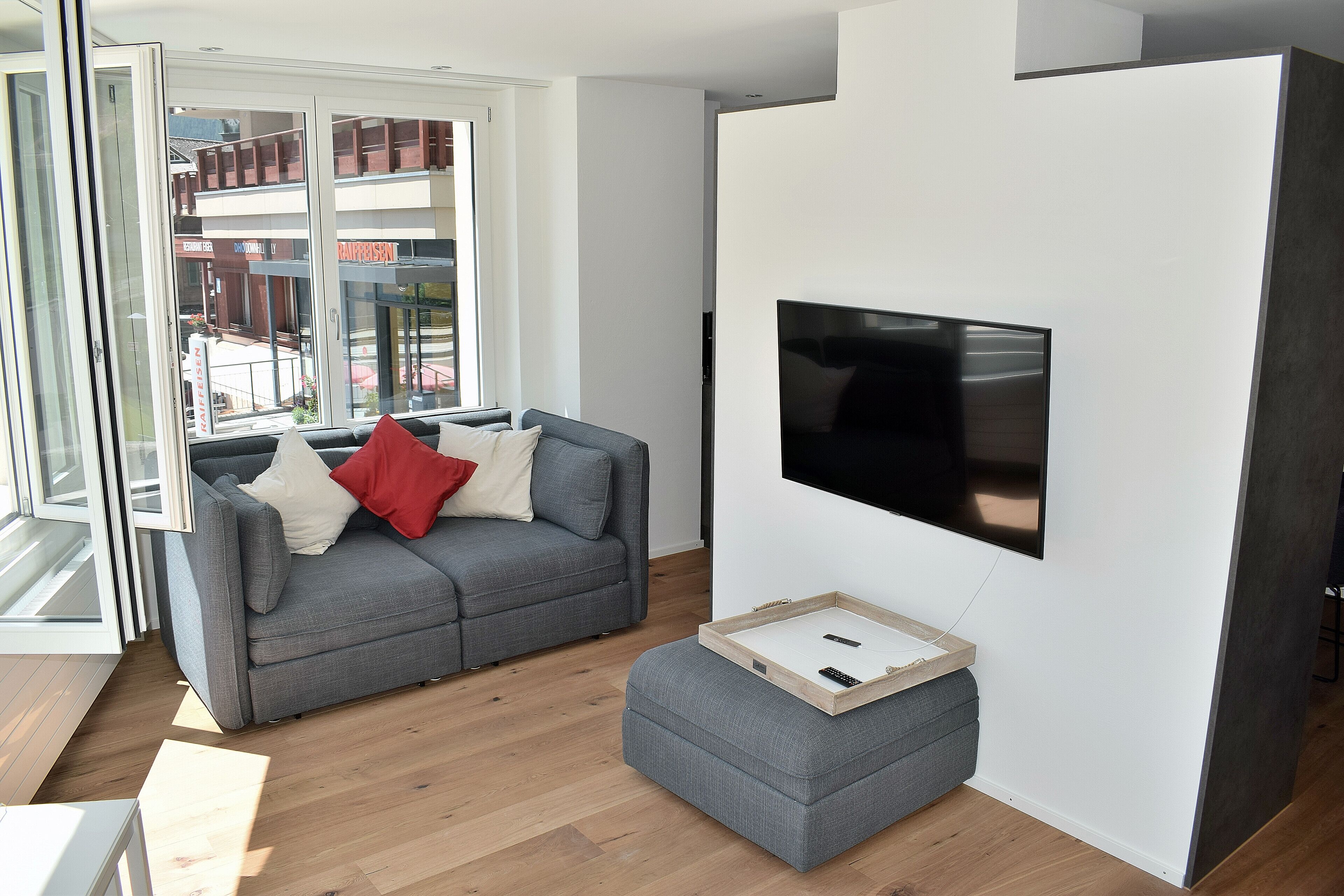 A modern living room with a grey sofa, a flat-screen TV mounted on the wall, and a wooden tray table.