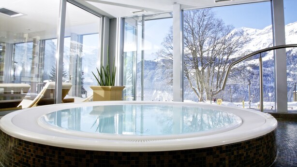 A modern indoor swimming pool with a panoramic mountain view, surrounded by large windows and furnished seating areas. - Hotel Silberhorn - Residences & Spa Wengen (Lauterbrunnen)