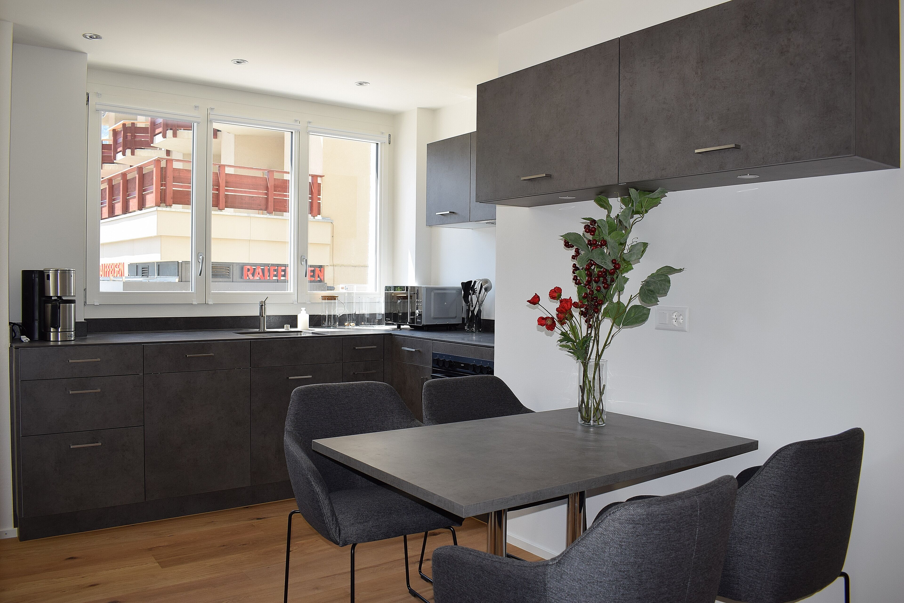 A modern kitchen with dark cabinets, a dining table with chairs, and a window with a view of a building.