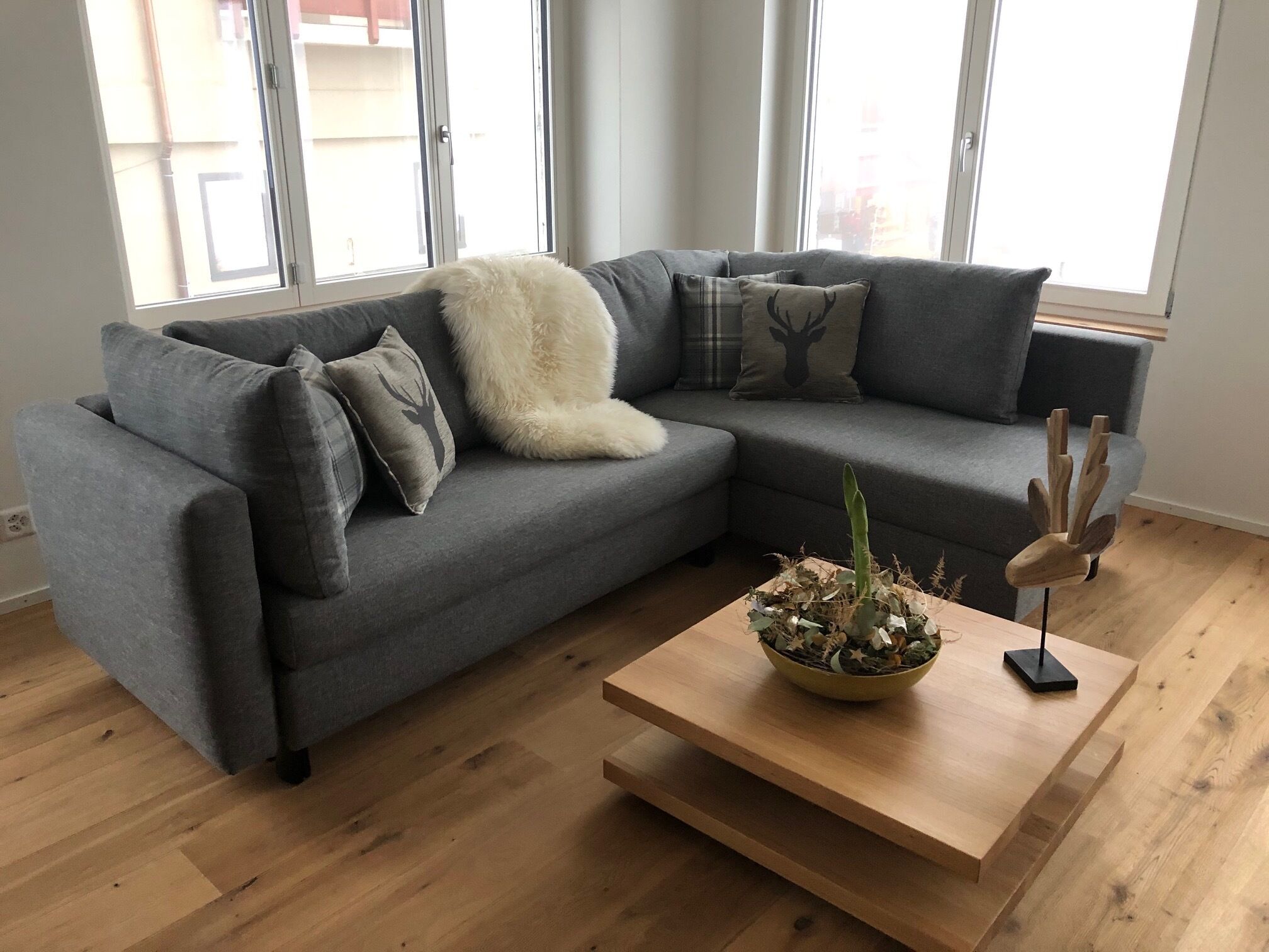 Un salon moderne avec un canapé d’angle gris, un plaid blanc et moelleux, et une table basse en bois agrémentée d’une plante décorative.