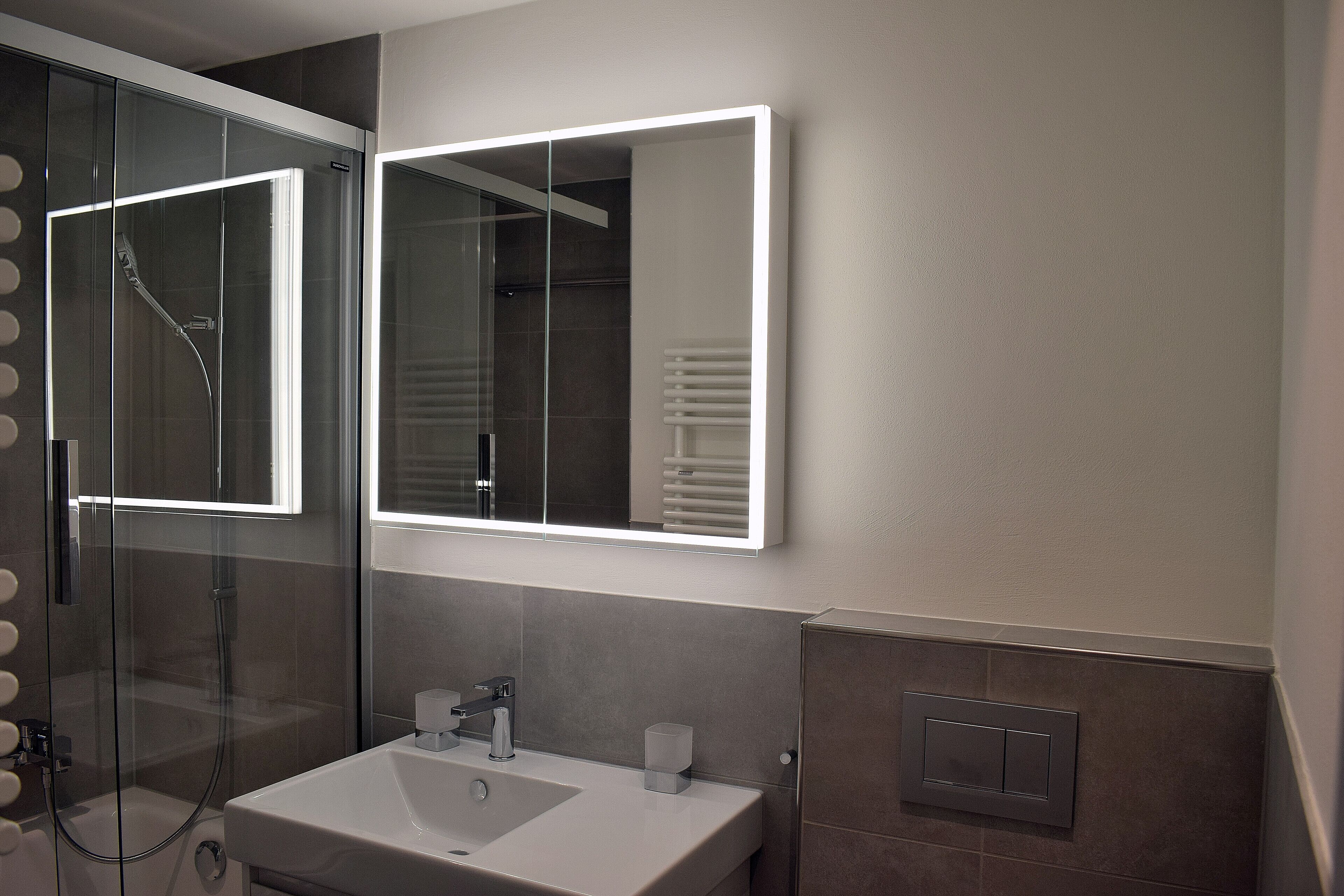 A modern bathroom with a glass-enclosed shower, a white sink, and a mirror with integrated lighting.