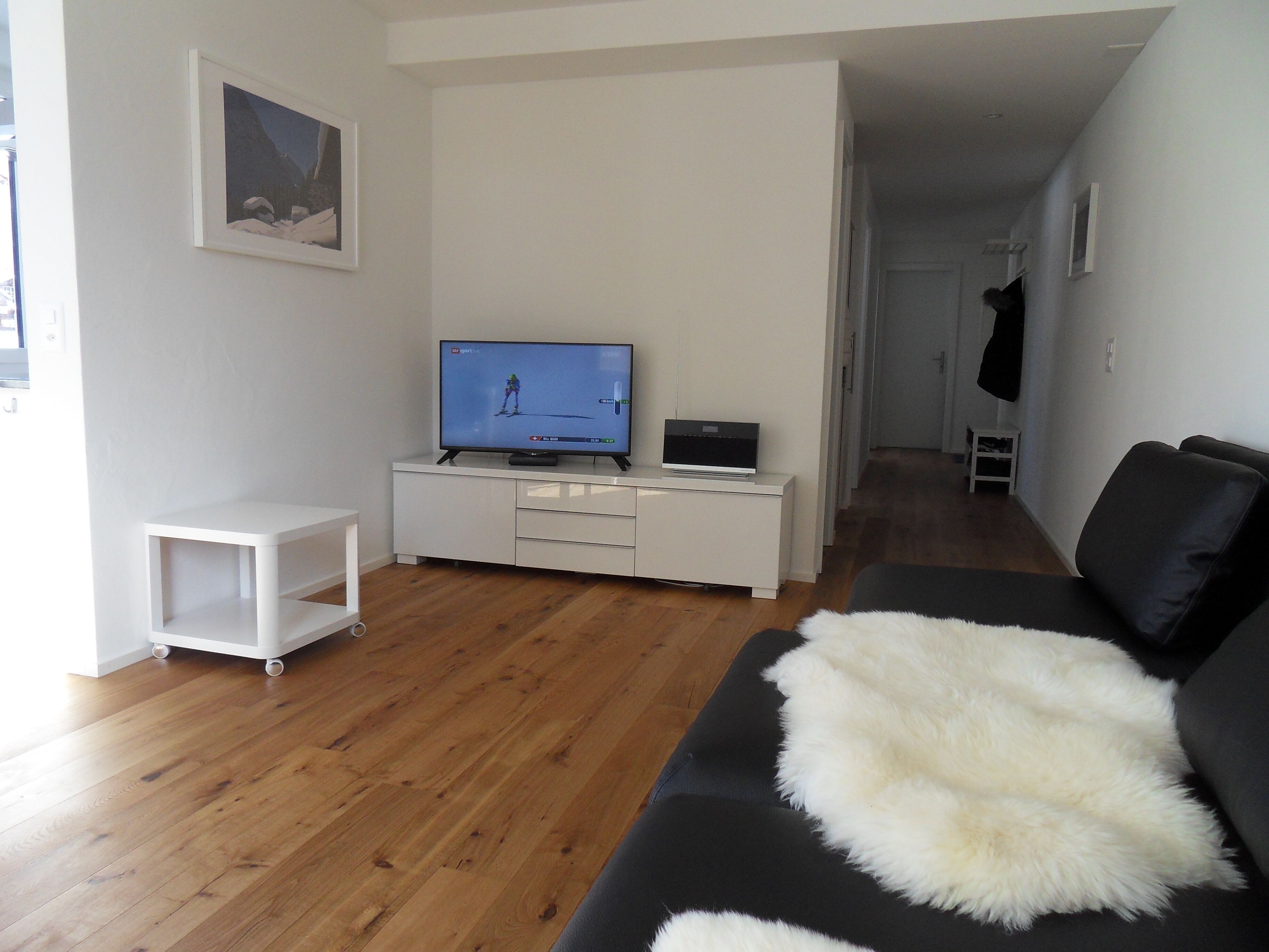 Ett modernt vardagsrum med en platt-TV på ett vitt skåp, en svart soffa med en vit fäll, trägolv och en inramad tavla på väggen.