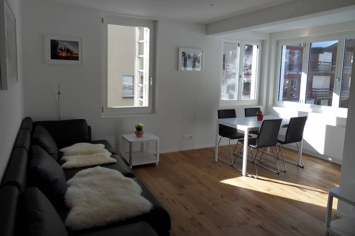 Un salon moderne avec un canapé noir, une petite table d’appoint blanche et un coin repas comprenant une table et des chaises blanches.