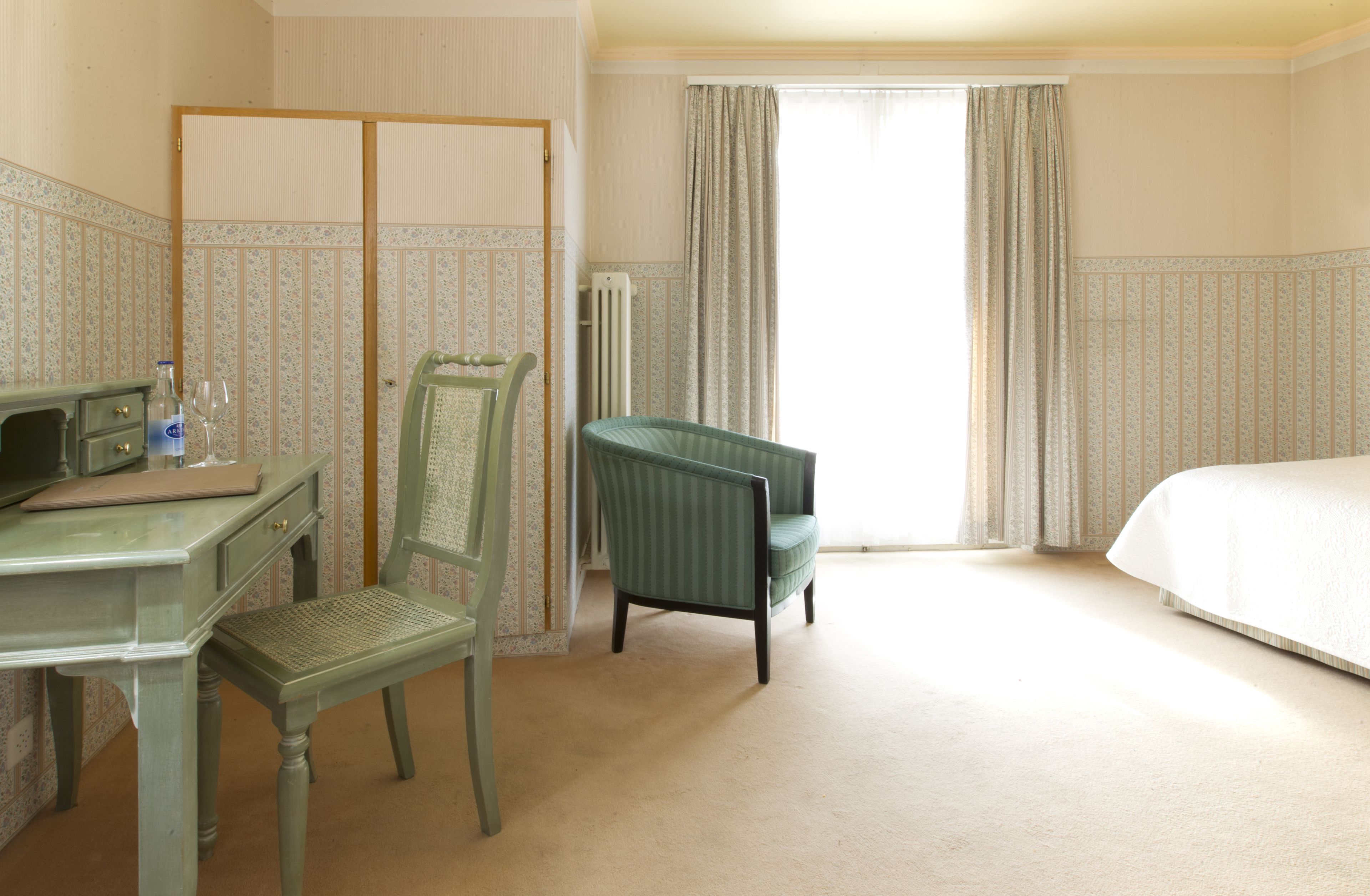 Une chambre d’hôtel avec un lit, un bureau et une chaise, un radiateur et une fenêtre avec des rideaux.