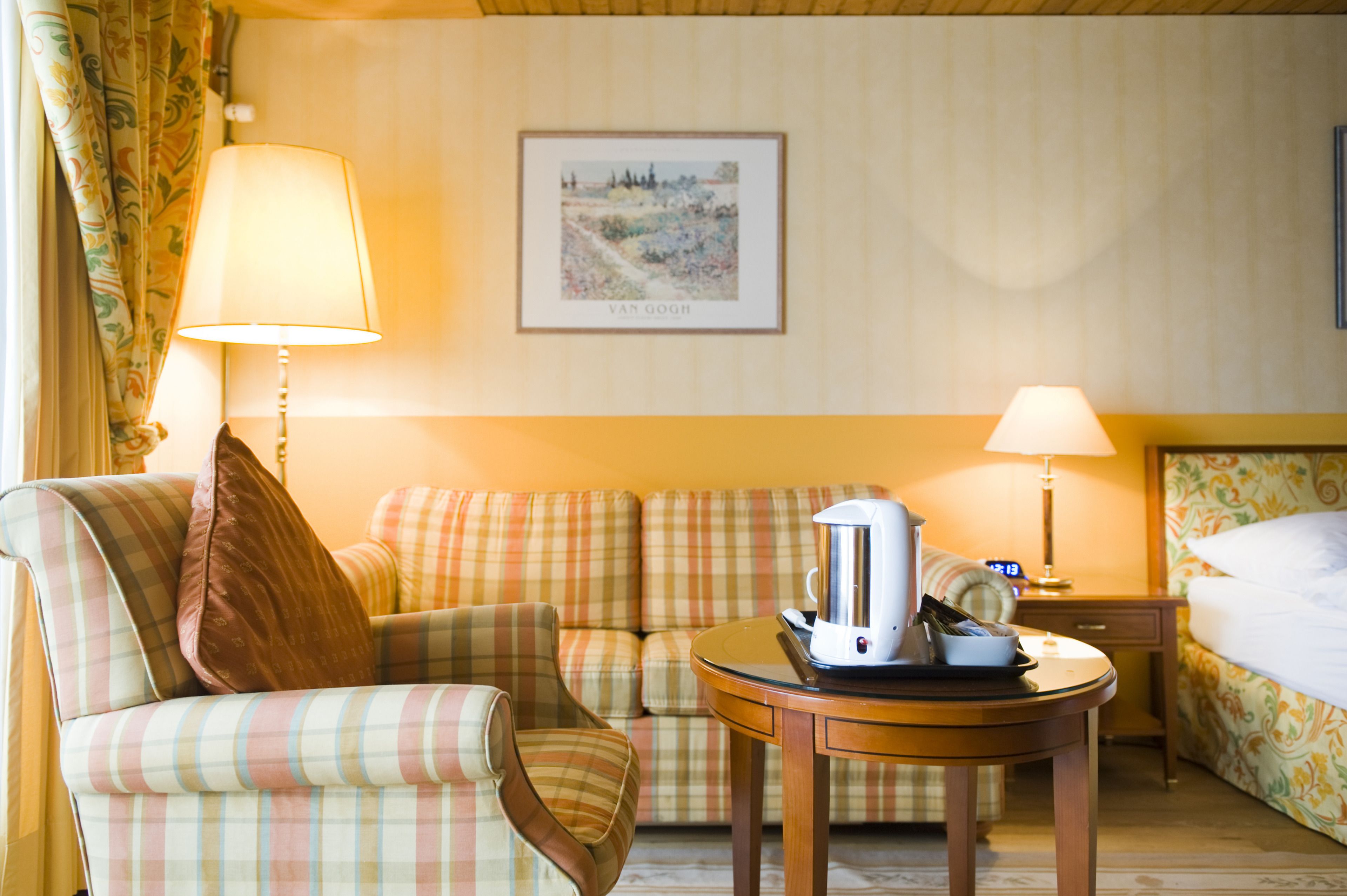 Habitación de hotel con sofá a cuadros, una pequeña mesa redonda con cafetera, cama con cabecera floral y un cuadro enmarcado en la pared.