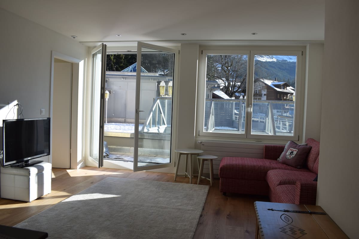 Un salon comprenant un téléviseur, un canapé, une table basse et une grande fenêtre donnant sur des maisons et des montagnes.