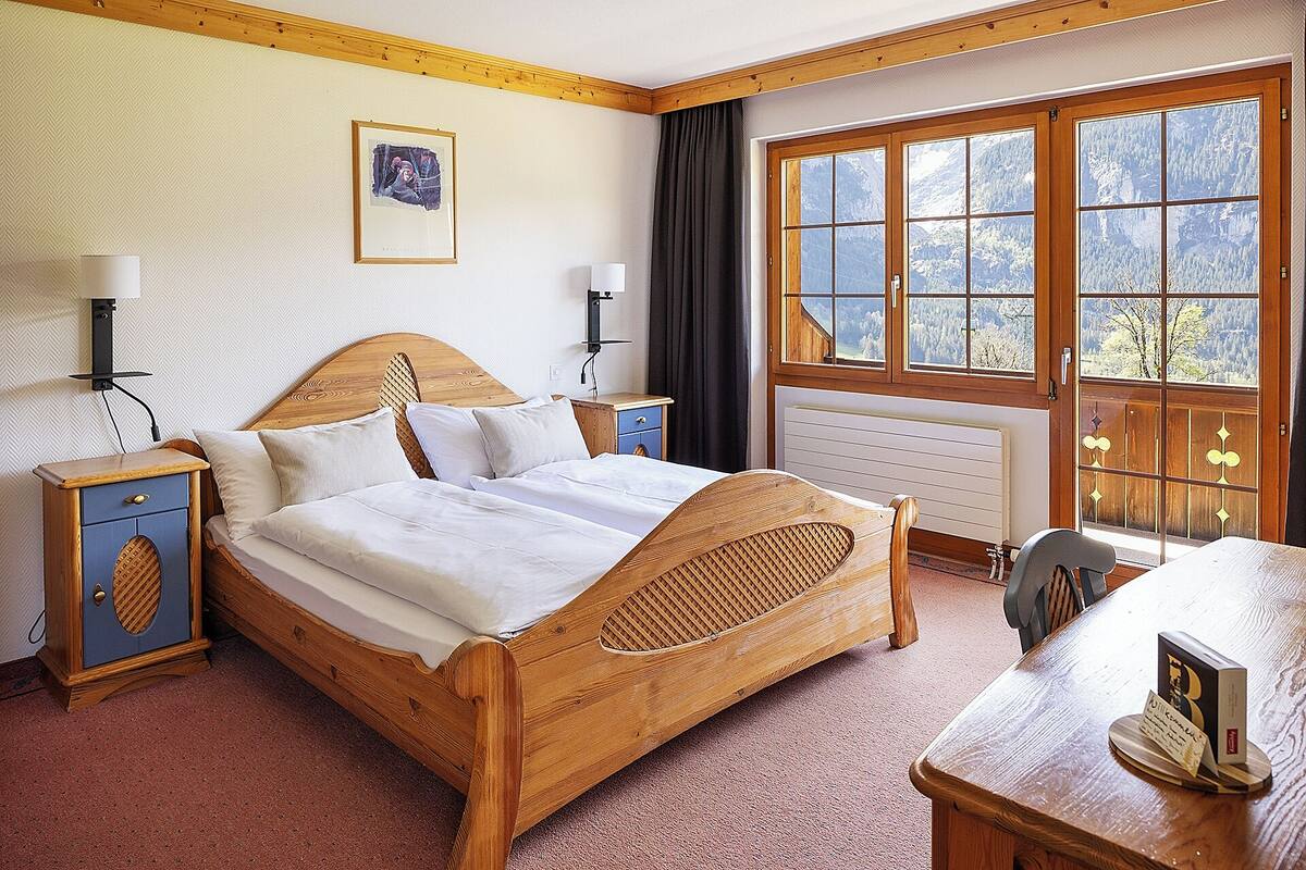 A wooden bed with a headboard, a bedside table, a lamp, and a window with a view of mountains.