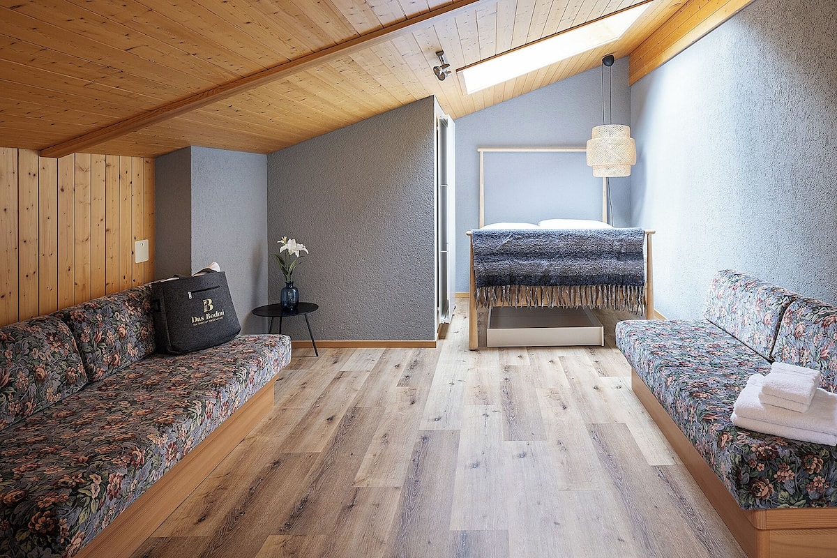 A modern room with a sloped ceiling, wooden flooring, a bed with a blue blanket, a sofa with a floral pattern, and a small table with a vase of flowers.