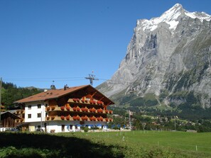 Un albergue de montaña con techo rojo, balcones adornados con macetas de flores y un teleférico al fondo.
