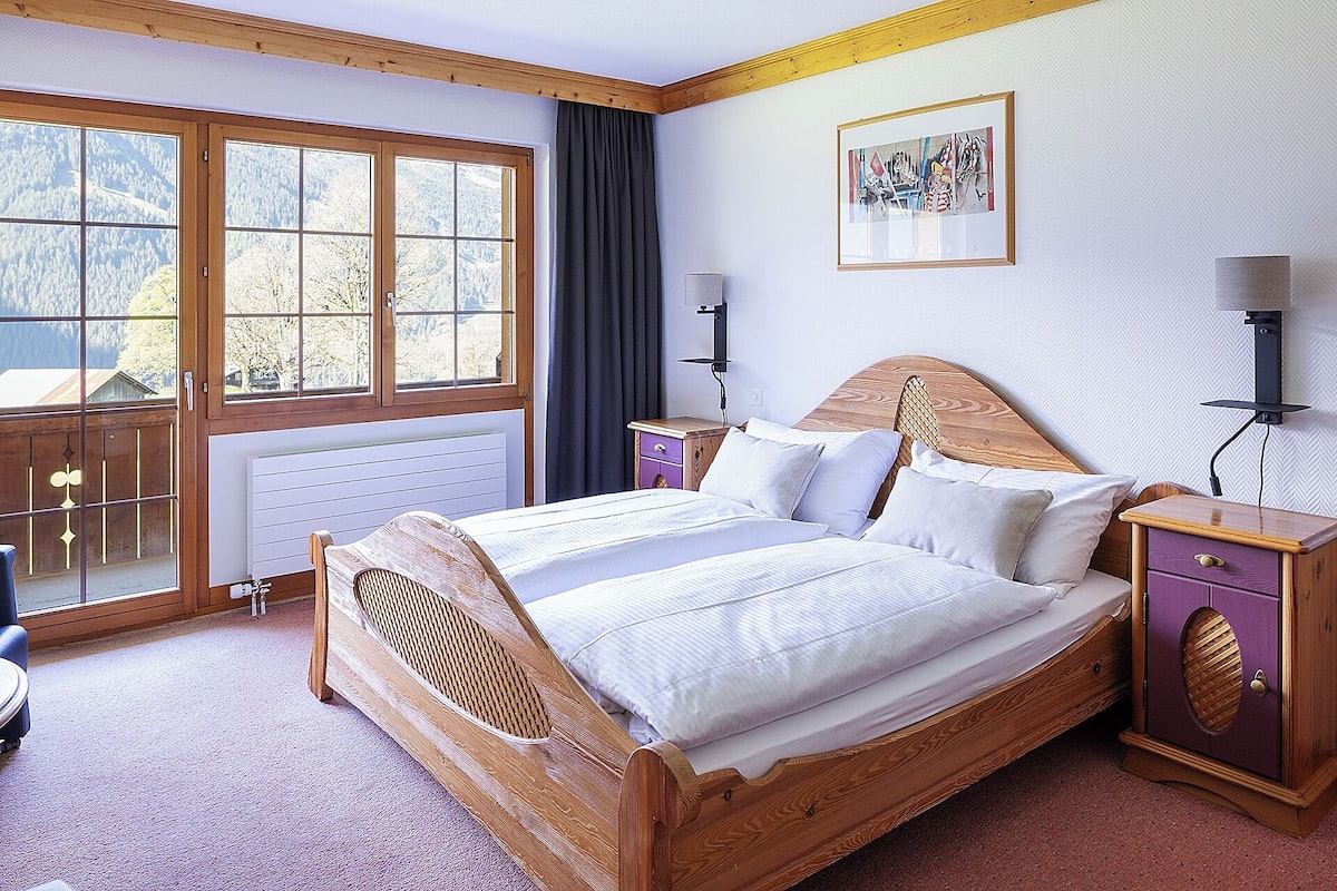 A wooden bed with a headboard, a nightstand, a lamp, and a window with a view of trees and mountains.