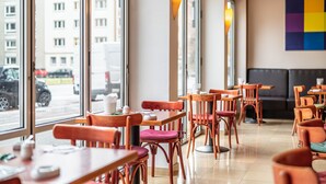 Un restaurant amb taules i cadires de fusta, grans finestres que donen al carrer i una obra d'art colorida a la paret.