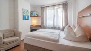 Un dormitori amb un llit, una cadira, un escriptori amb una làmpada, una televisió i una finestra amb cortines.