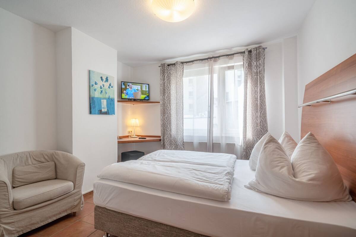 Une chambre à coucher comprenant un lit, une chaise, un bureau avec une lampe, un téléviseur et une fenêtre avec des rideaux.